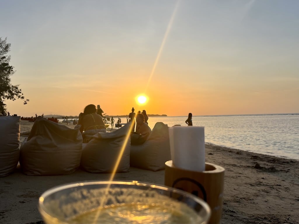 ギリ・アイルのビーチバーから見る海に沈む夕日。砂の上のビーズクッションと、水平線に浮かぶギリ・メノのシルエット