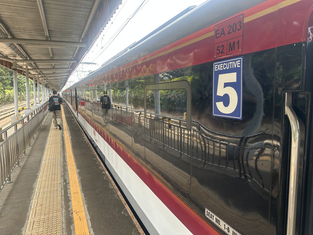 Trem do aeroporto vermelho, branco e preto parado numa plataforma ao ar livre, com um vagão da classe Executive número 5 em primeiro plano
