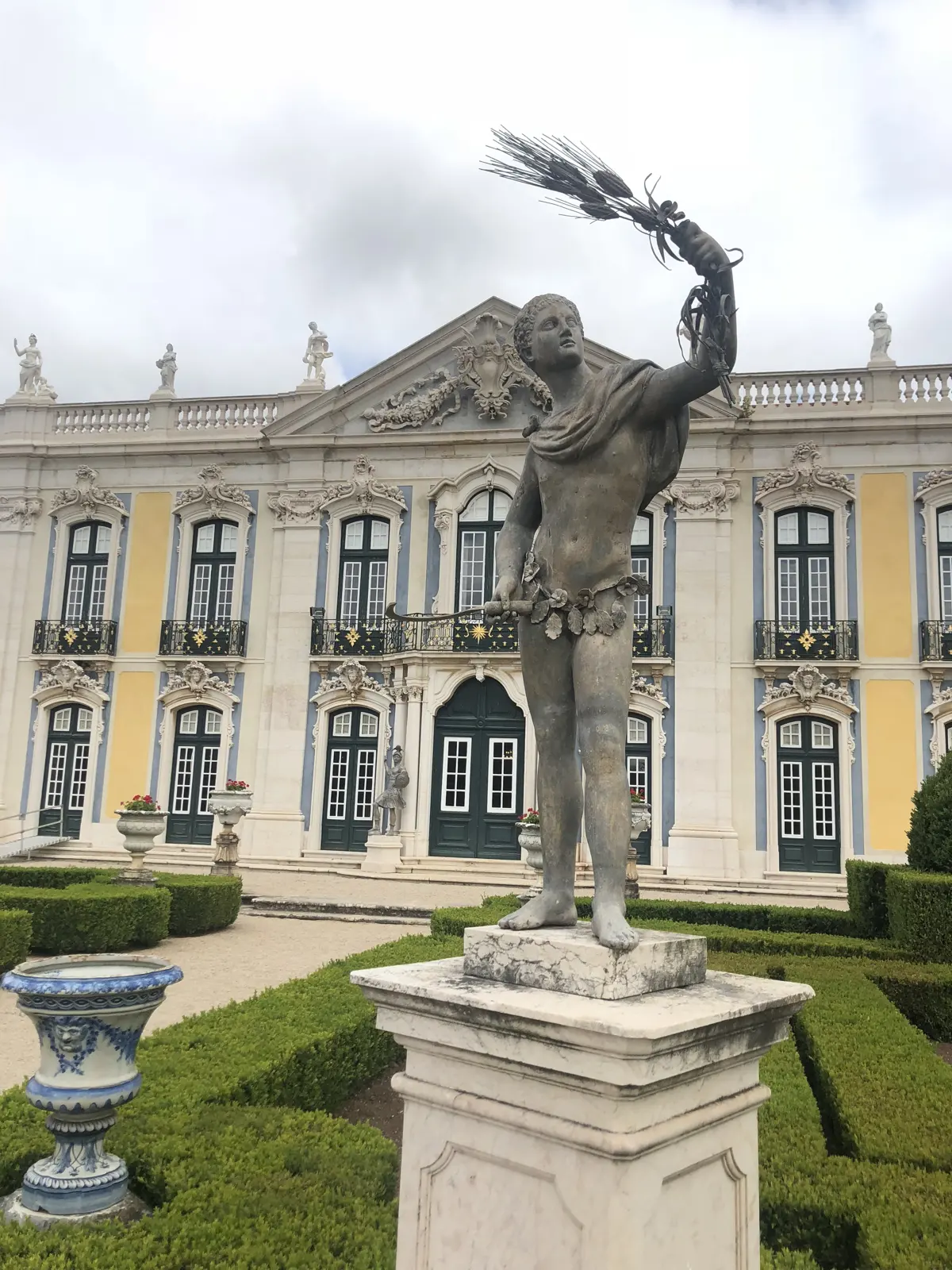A statue in the formal gardens in front of the yellow facade of the Palace of Queluz